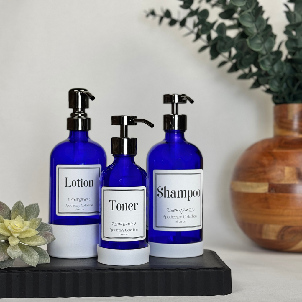 Three cobalt blue glass dispensers with white silicone sleeves and Apothecary labels for lotion, toner and shampoo